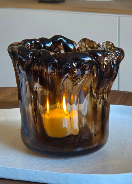 Waxinelichthouder Myron in donkerbruin glas met golvende druipende rand en brandend waxinelichtje op een witte ondergrond