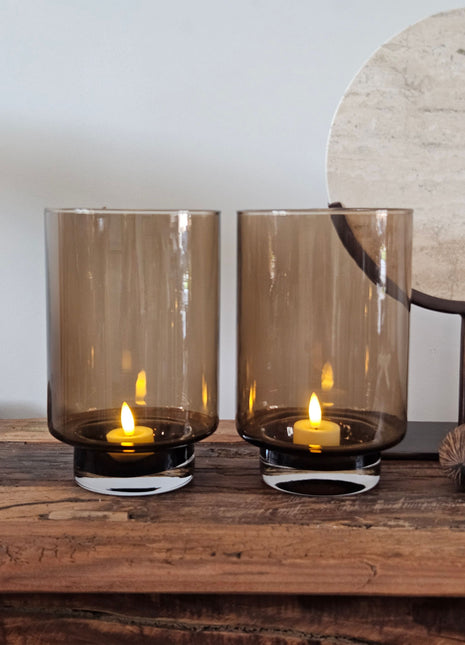 Twee cilindrische windlichten Dune Taupe S in rookglas met LED-kaarsen naast een rond travertijn ornament op een zwarte standaard en twee droge noten op een houten ondergrond