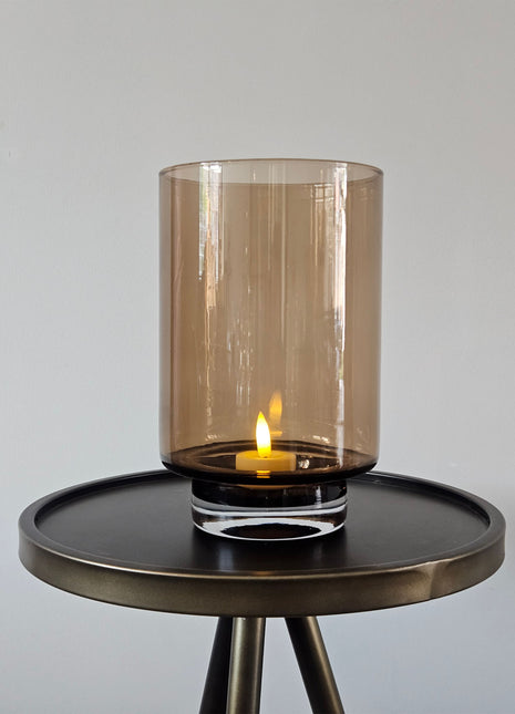 Cilindrisch windlicht Dune Taupe S in Taupe met een dikke glazen bodem en een brandende LED-kaars op een rond bronzen bijzettafeltje